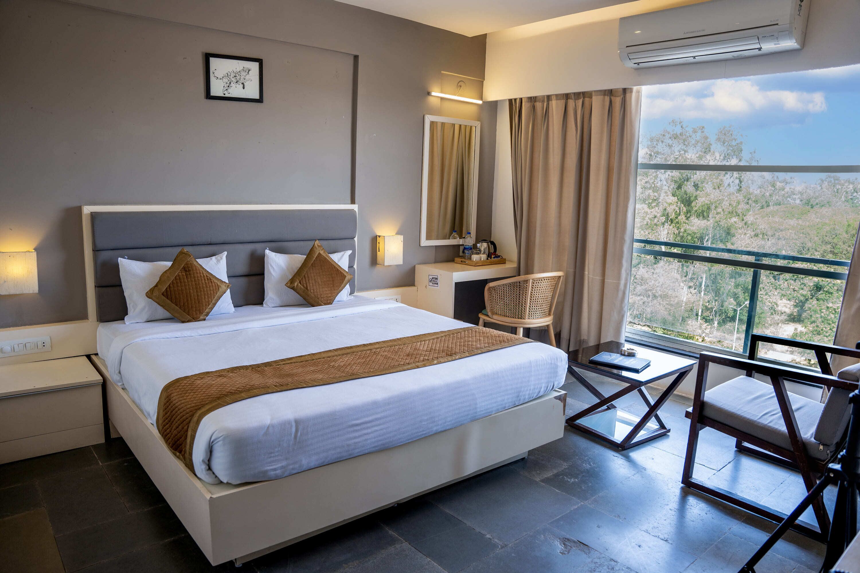 Quarto de hotel moderno com uma cama grande, área de estar com uma poltrona, uma pequena mesa e varanda com vista para árvores.