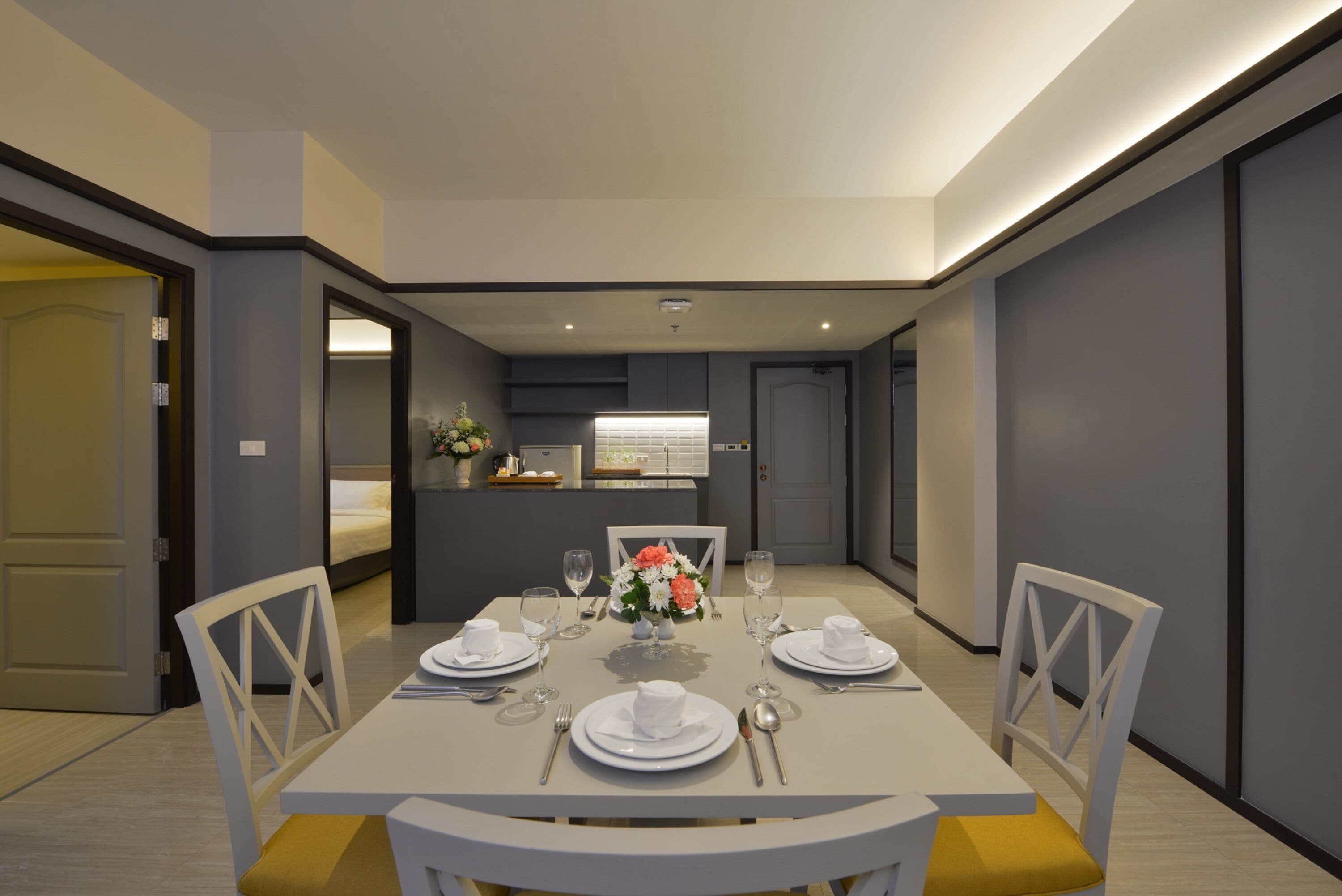 Un comedor moderno con una mesa blanca para dos, un jarrón de flores y vista a una cocina.