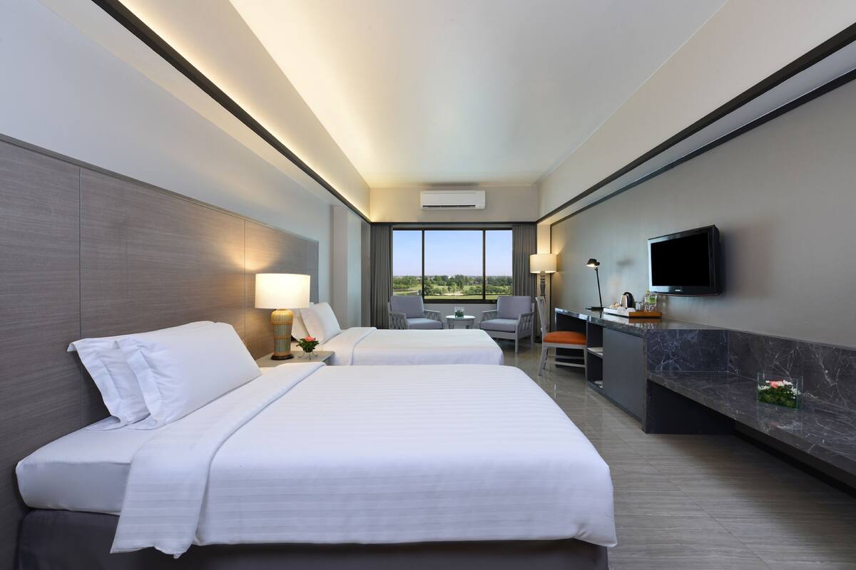 A hotel room with two beds, a desk, a chair, a TV, and a view of the outdoors.