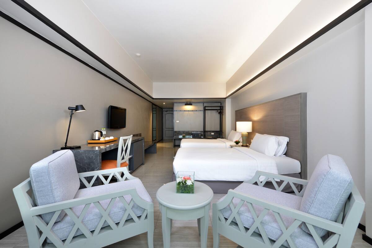 A hotel room with a bed, two armchairs, a small table, a desk with a lamp, a TV, and a bathroom in the background.
