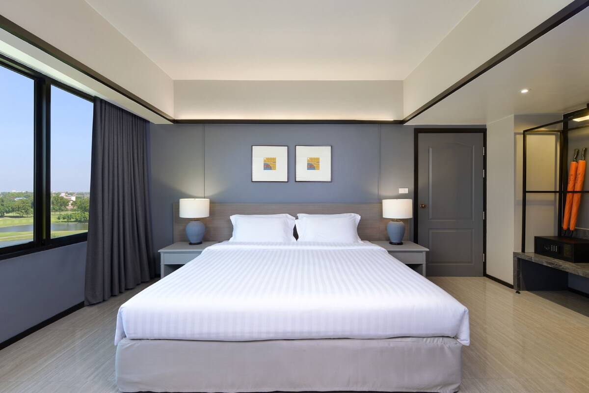 A modern hotel room with a large bed, two bedside tables with lamps, a view of the outdoors, and a door leading to a bathroom.