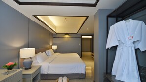 A hotel room with a neatly made bed, a bedside table with a lamp, a robe hanging on a rack, and a small basket with slippers. - Tinidee Hotel Bangkok Golf Club (Pathum Thani)