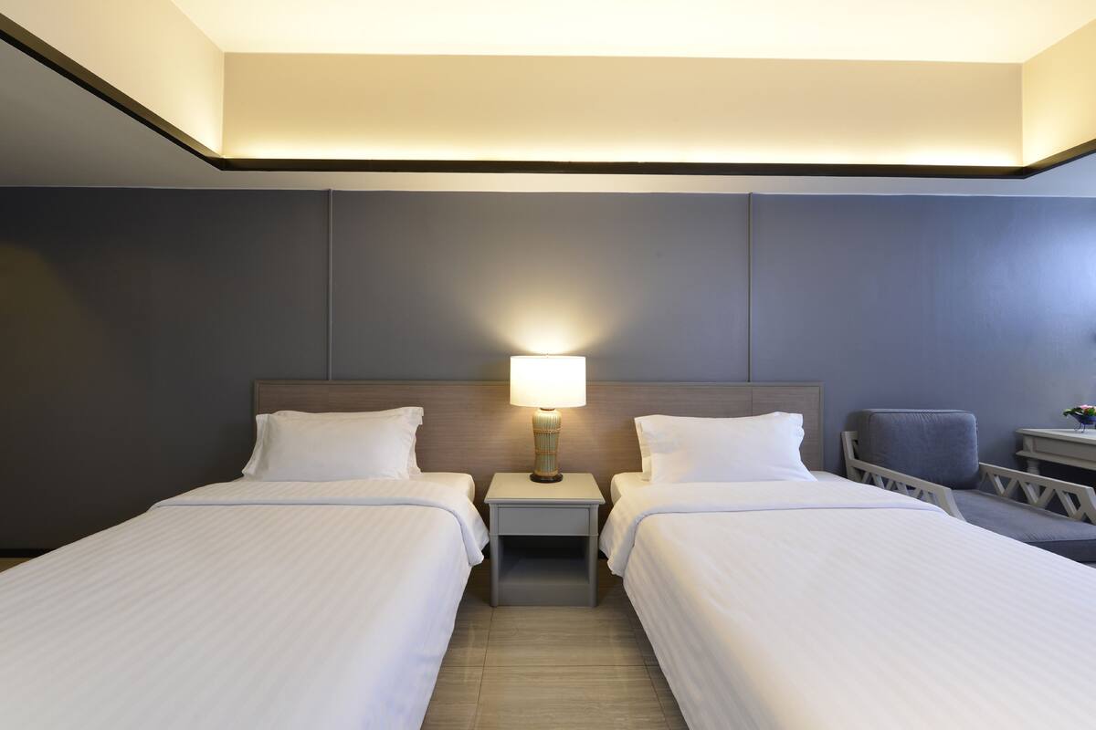 A hotel room with two beds, a nightstand with a lamp, a chair, and a small table with a vase.