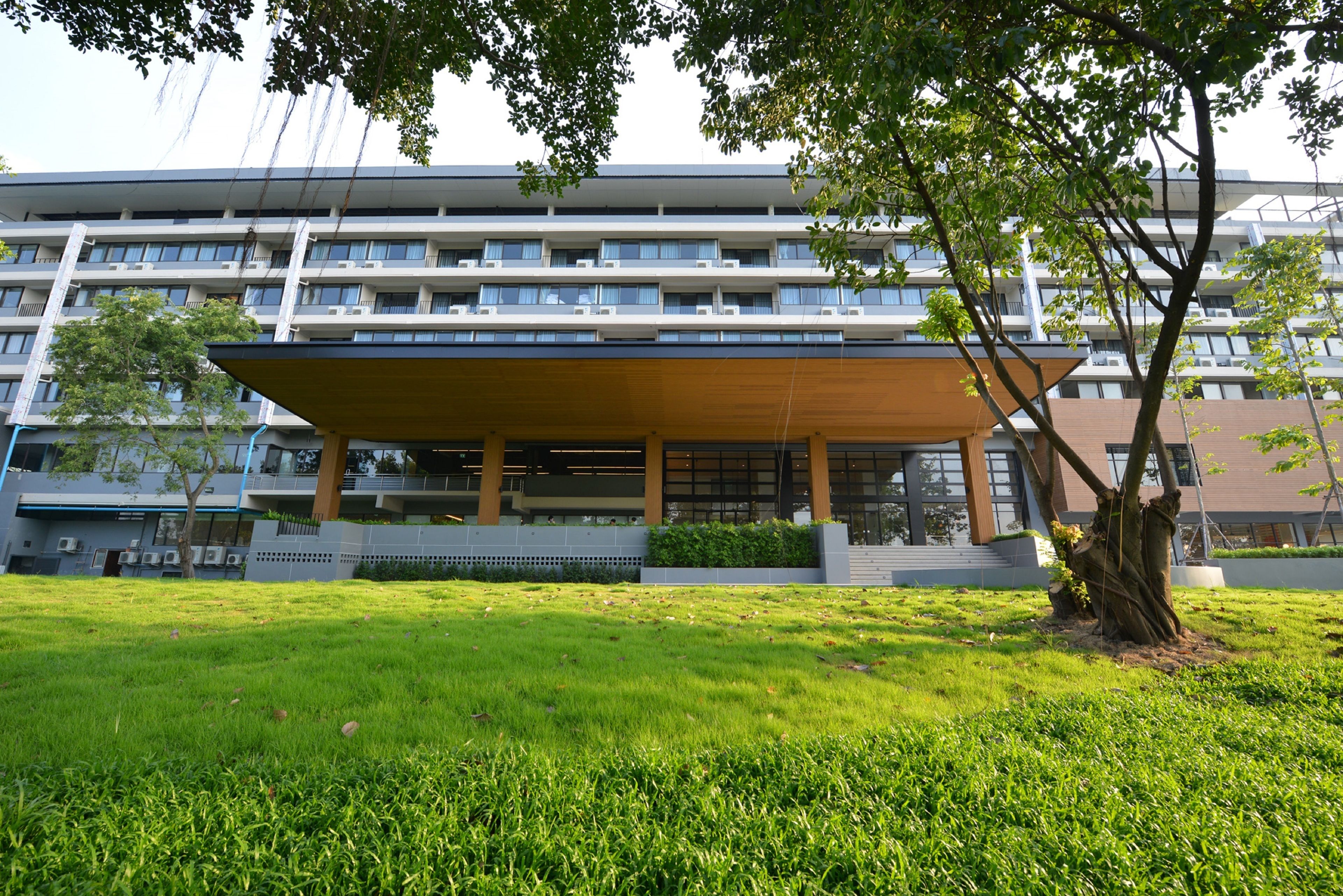 Un bâtiment moderne doté d’un espace extérieur couvert, entouré d’un champ verdoyant et d’arbres.