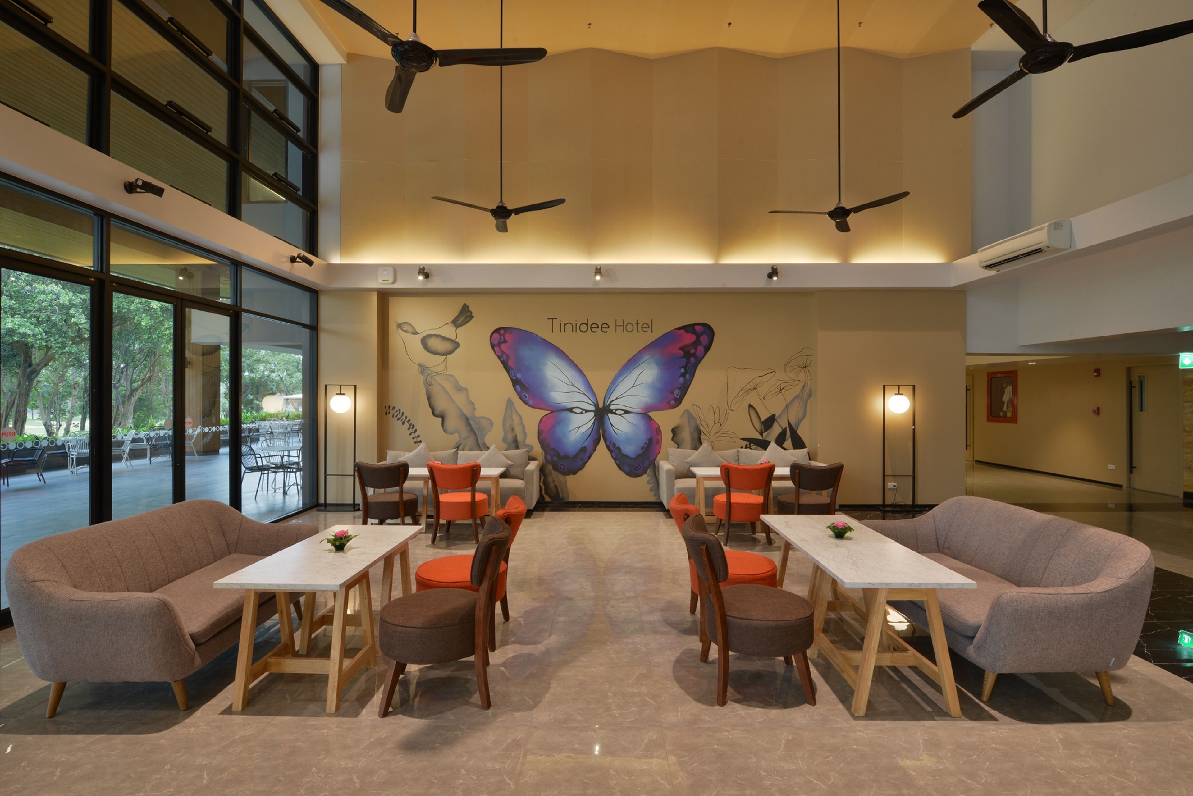 Un vestíbulo de hotel con un gran mural de mariposas, ventiladores de techo y un área de descanso con mesas y sillas.