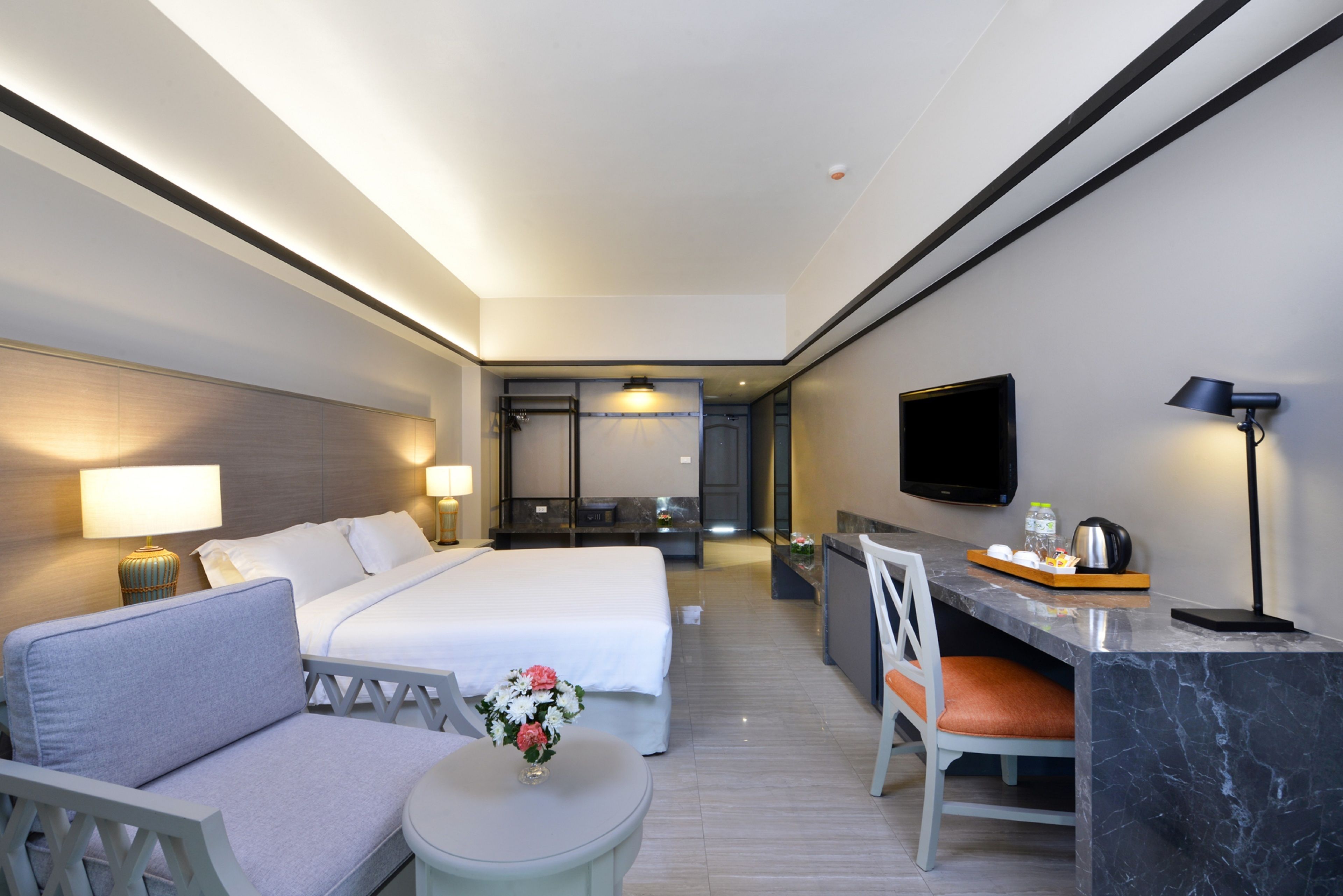 A hotel room with a bed, a desk, a chair, a small table, a TV, and a lamp.