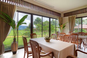 Comedor con una mesa dispuesta para seis personas, sillas de madera, ventanas amplias con vistas a zonas verdes y edificios, y un jarrón con flores sobre la mesa.