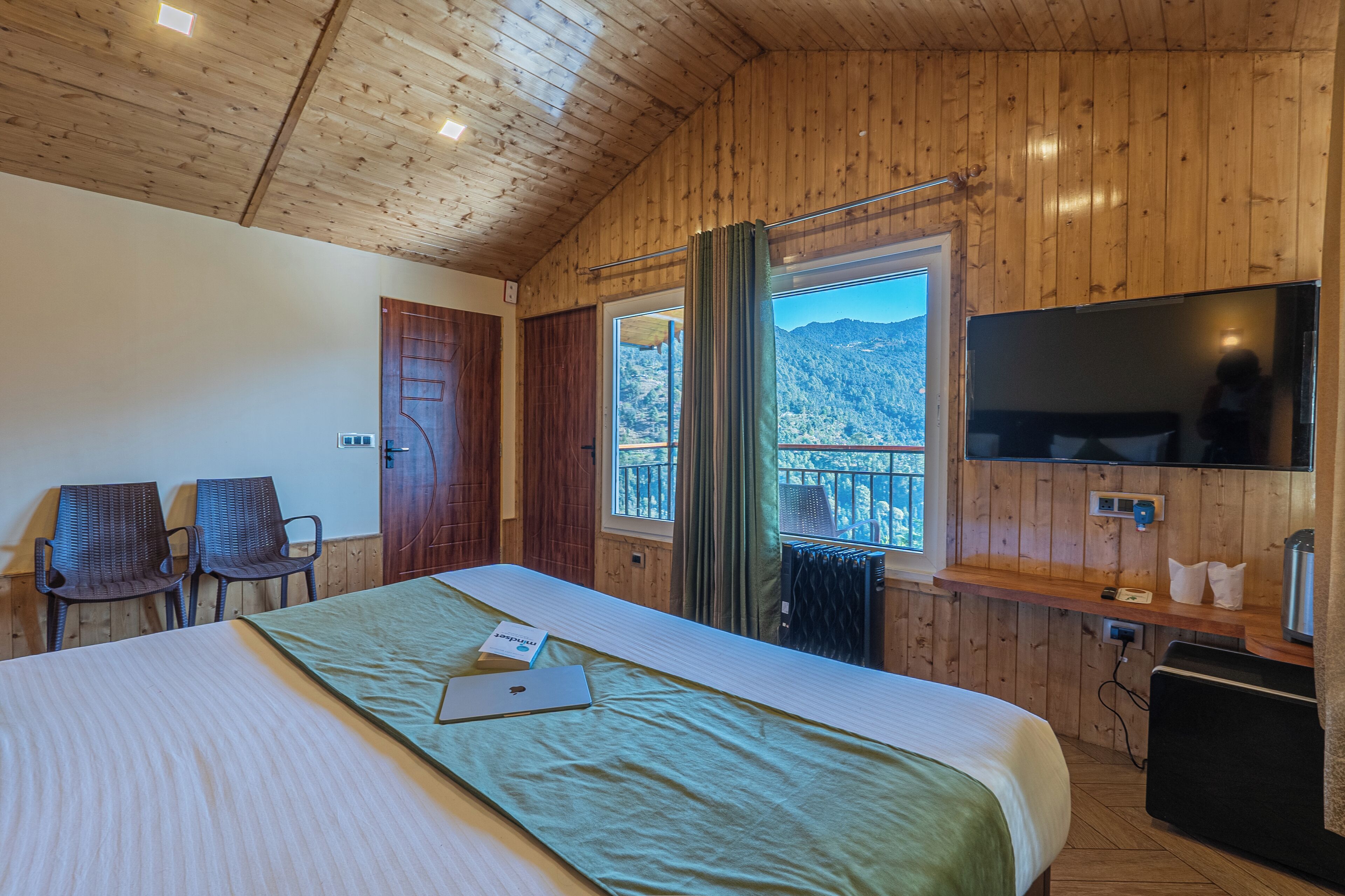 Una habitación con paneles de madera, una cama, dos sillas, un televisor y una ventana con vista a las montañas.