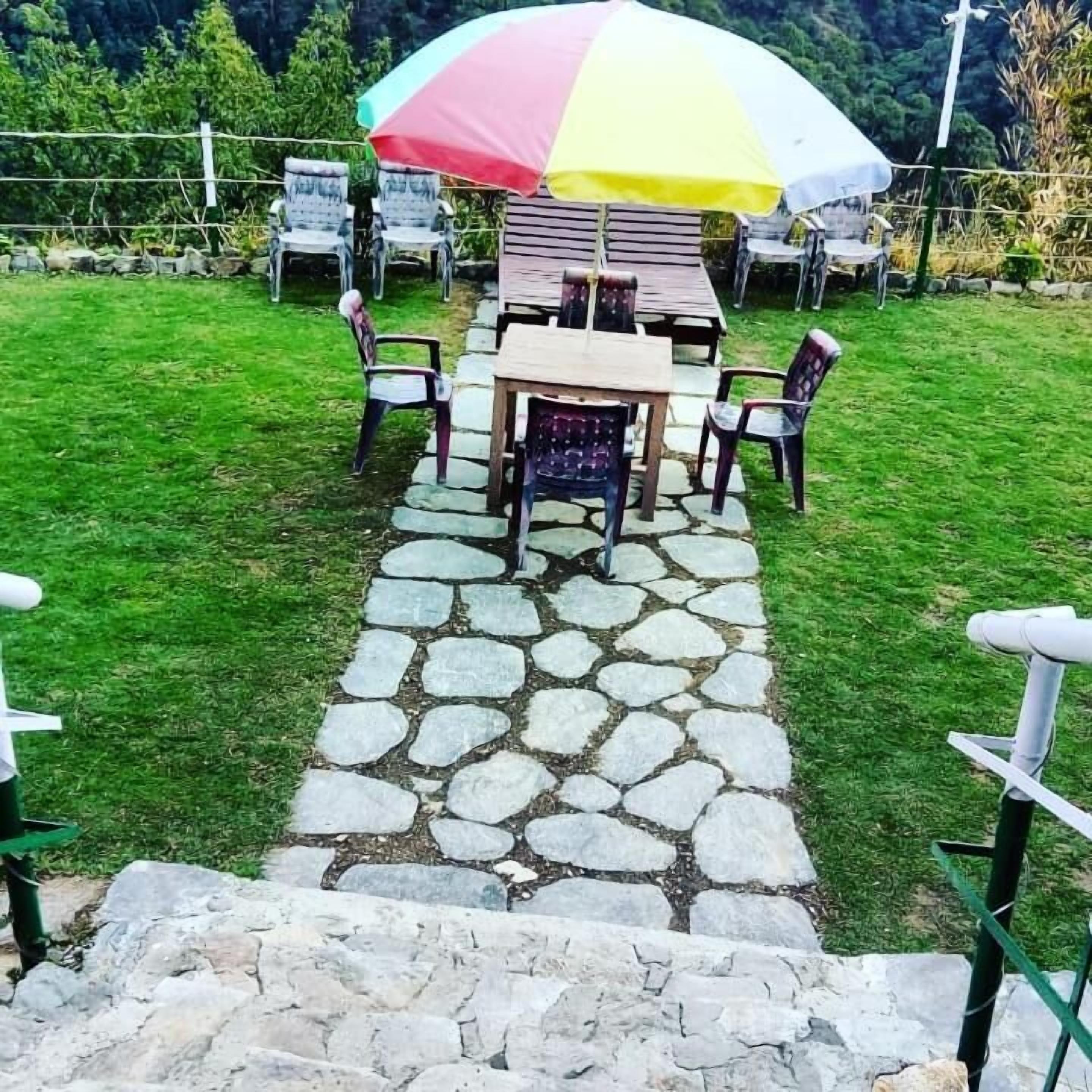 A stone pathway leading to a wooden table and chairs set up outdoors, surrounded by a grassy area and a stone wall.