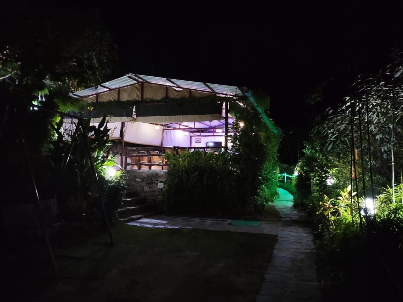 A covered outdoor seating area with tables and chairs, surrounded by greenery and a stone pathway.