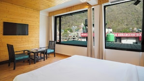 A hotel room with a wooden ceiling, a bed, a TV mounted on the wall, a table with chairs, and large windows offering a view of buildings and a water tank outside. - Hotel Ishan A Riverside Retreat (Narendranagar)
