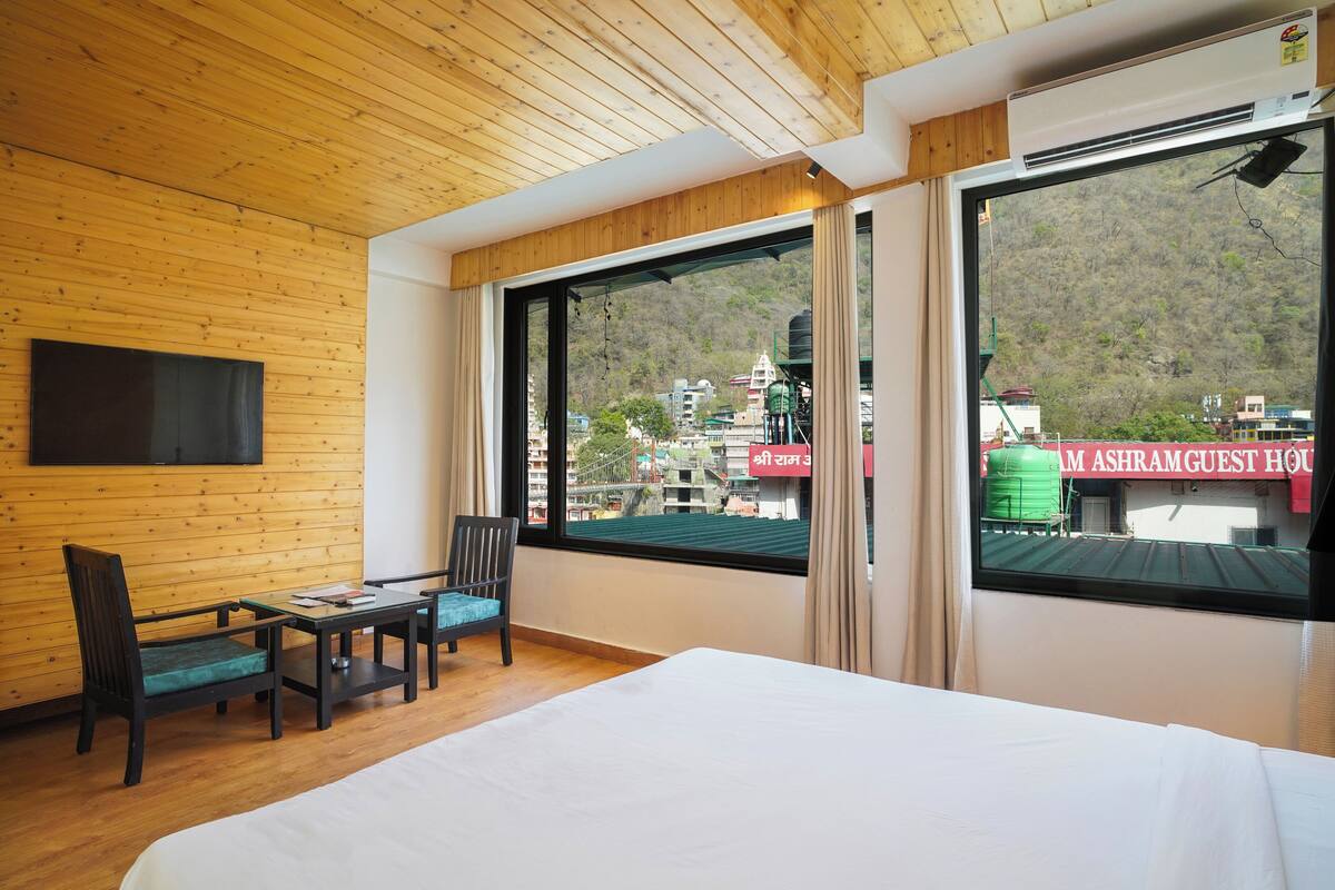 A hotel room with a wooden ceiling, a bed, a TV mounted on the wall, a table with chairs, and large windows offering a view of buildings and a water tank outside.