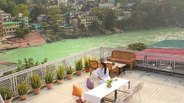 A terrace with a table and chairs, overlooking a river and a hillside of colorful buildings. - Hotel Ishan A Riverside Retreat (Narendranagar)