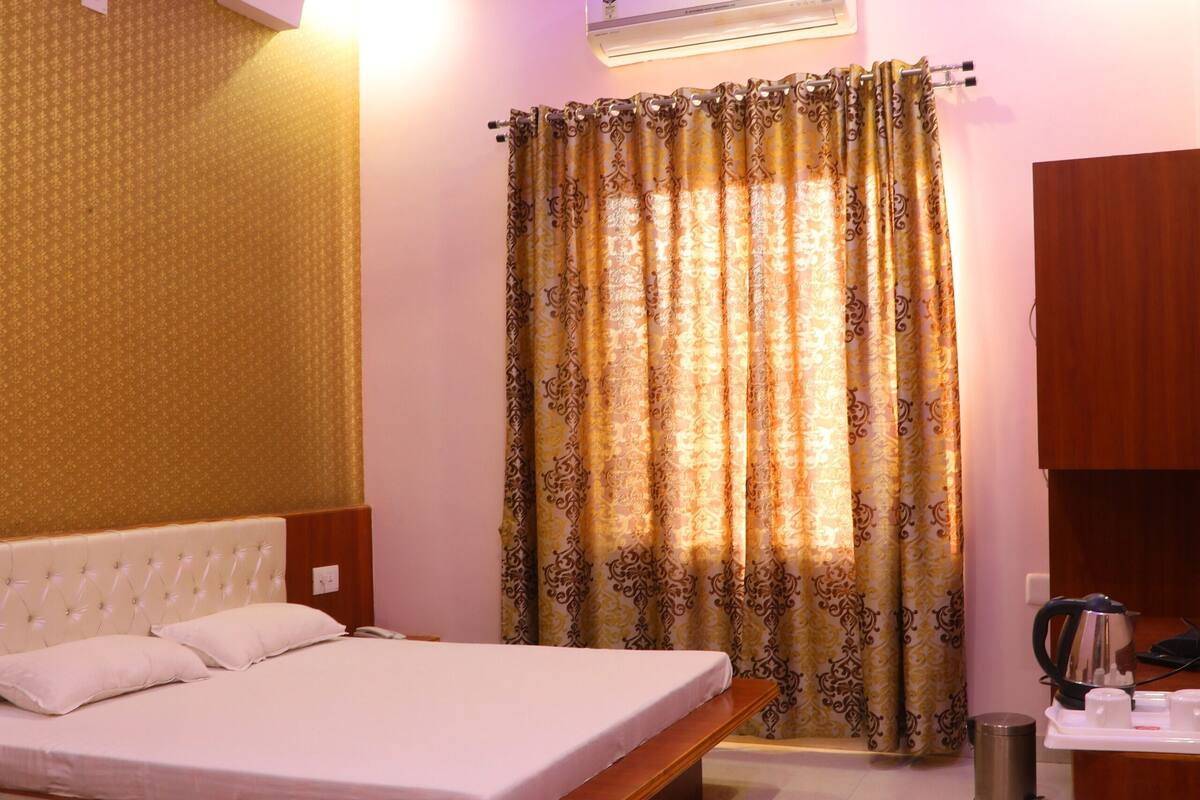 A bedroom with a bed, a ceiling fan, an air conditioning unit, a bedside table, a desk, and a curtain.
