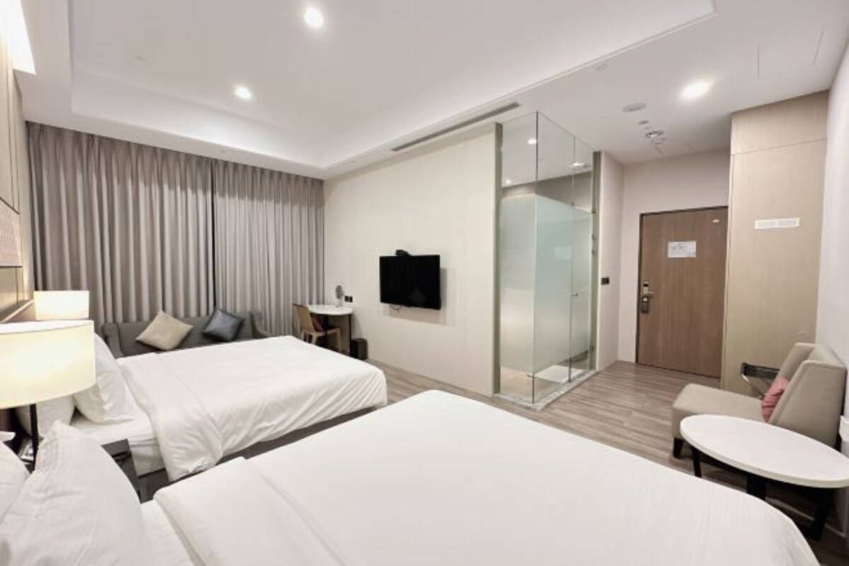 A hotel room with two beds, a TV, a desk, and a glass partition.