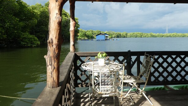 A wooden deck with a metal table and chair, overlooking a body of water with a blue building in the distance. - Bakau Hijau Riverlodge - Hostel (Bedung)