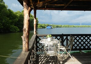 A wooden deck with a metal table and chair, overlooking a body of water with a blue building in the distance. - Bakau Hijau Riverlodge - Hostel (Bedung)