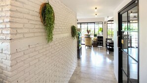 A hallway with white brick walls, hanging plants, and a glass door leading to a seating area with tables and chairs.