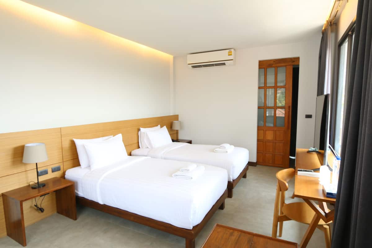 A hotel room with two beds, a desk, a chair, a lamp, an air conditioning unit, and a window with curtains.