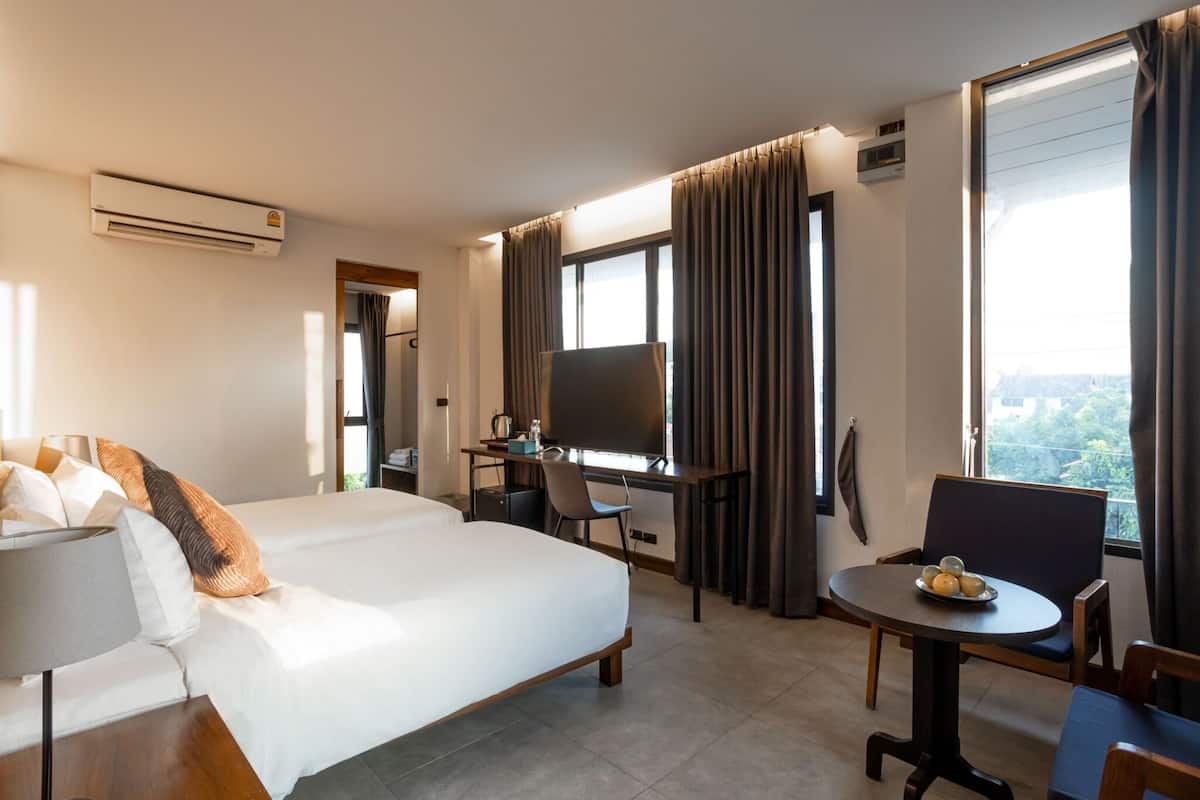 A hotel room with a bed, a desk with a computer, a chair, a small table with a bowl of fruit, and large windows with curtains.