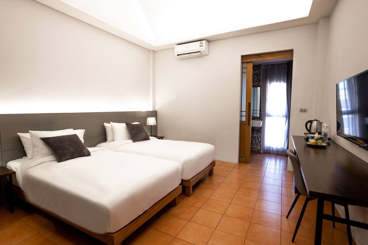 A hotel room with two beds, a TV, a dining table, and an air conditioning unit.