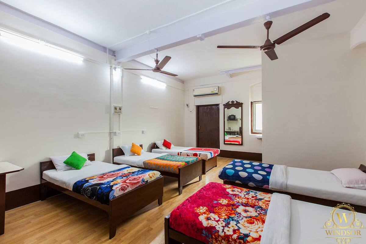 A dormitory room with four single beds, each with colorful bedding. There are ceiling fans, a large mirror, and an air conditioning unit on the wall.