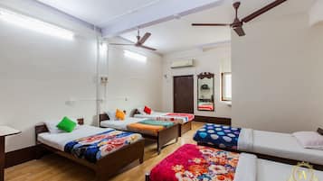 A dormitory room with four single beds, each with colorful bedding. There are ceiling fans, a large mirror, and an air conditioning unit on the wall.