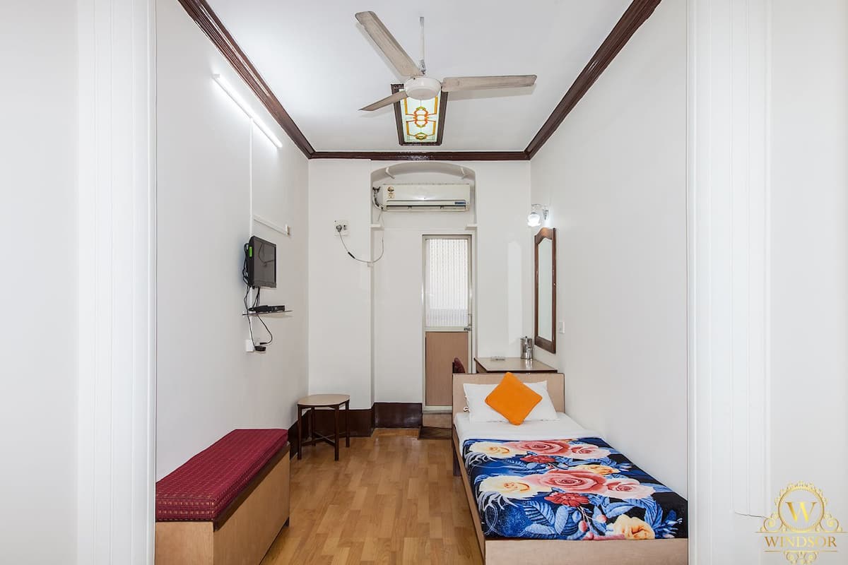 A compact hotel room with a bed, a desk, a chair, a ceiling fan, an air conditioning unit, and a television.