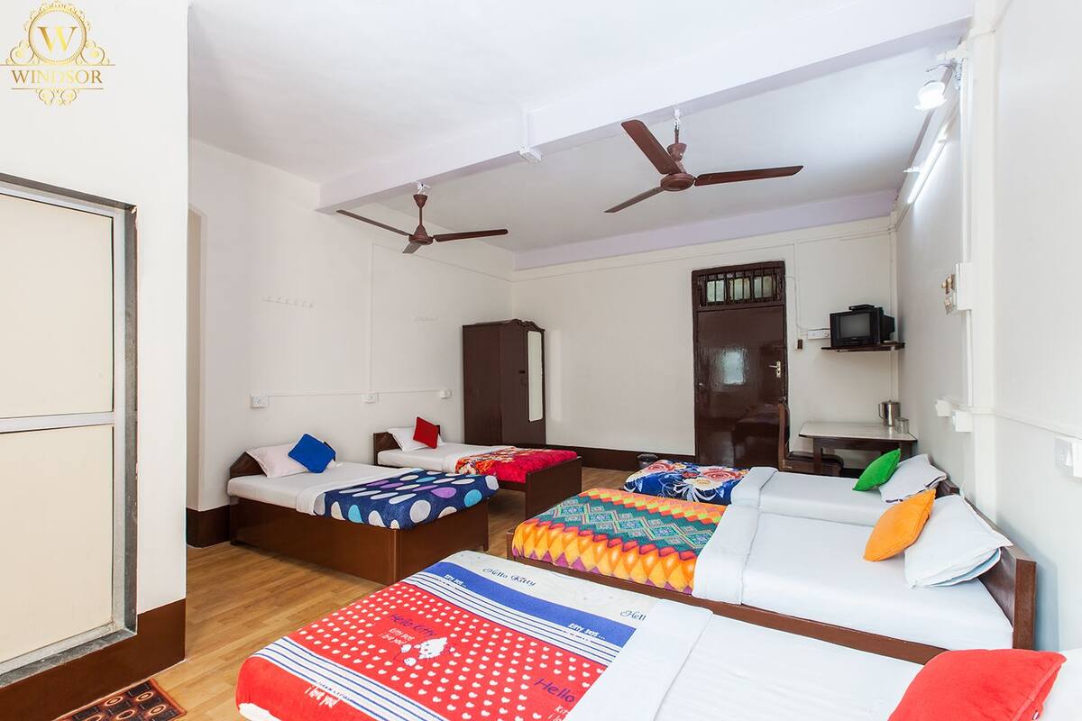 A hotel room with three beds, a wardrobe, a desk with a television, and ceiling fans.