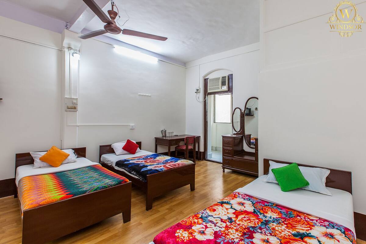 A room with three wooden beds, each with colorful bedding. There is a ceiling fan, a desk with a chair, and a mirror on the wall.