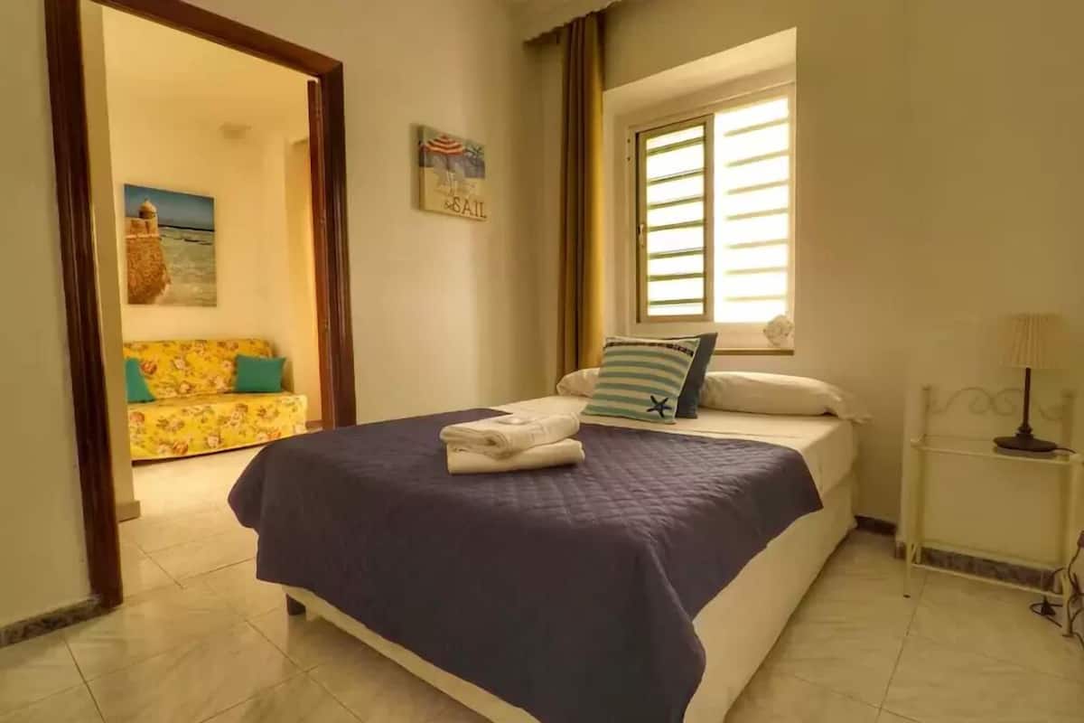 A bedroom with a bed, a window with blinds, a painting on the wall, and a door leading to another room.