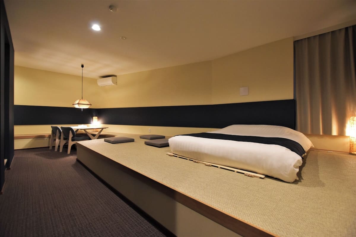 A hotel room with a bed, a desk, a chair, a hanging light fixture, and a window with curtains.