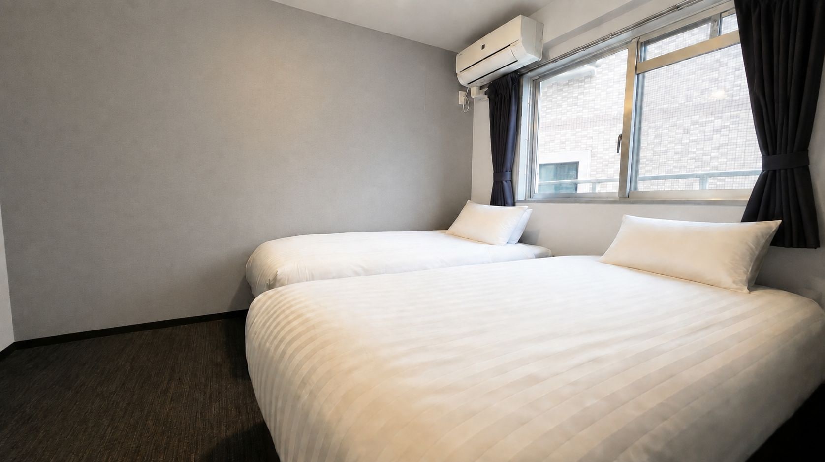 A hotel room with two beds, a window with curtains, and an air conditioning unit.