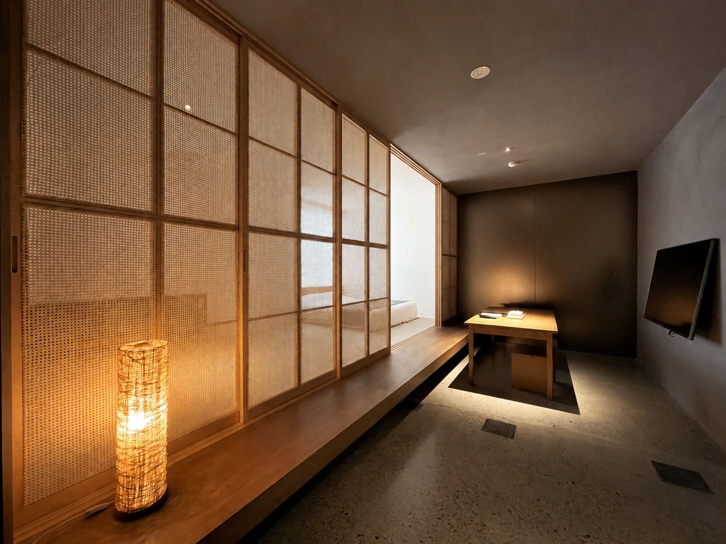 A room with a tatami mat floor, a low wooden table, a wall-mounted TV, and a traditional Japanese paper lantern.