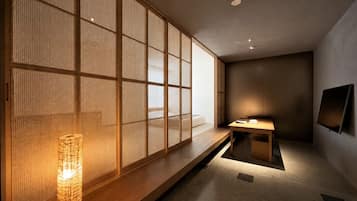 A room with a tatami mat floor, a low wooden table, a wall-mounted TV, and a traditional Japanese paper lantern.