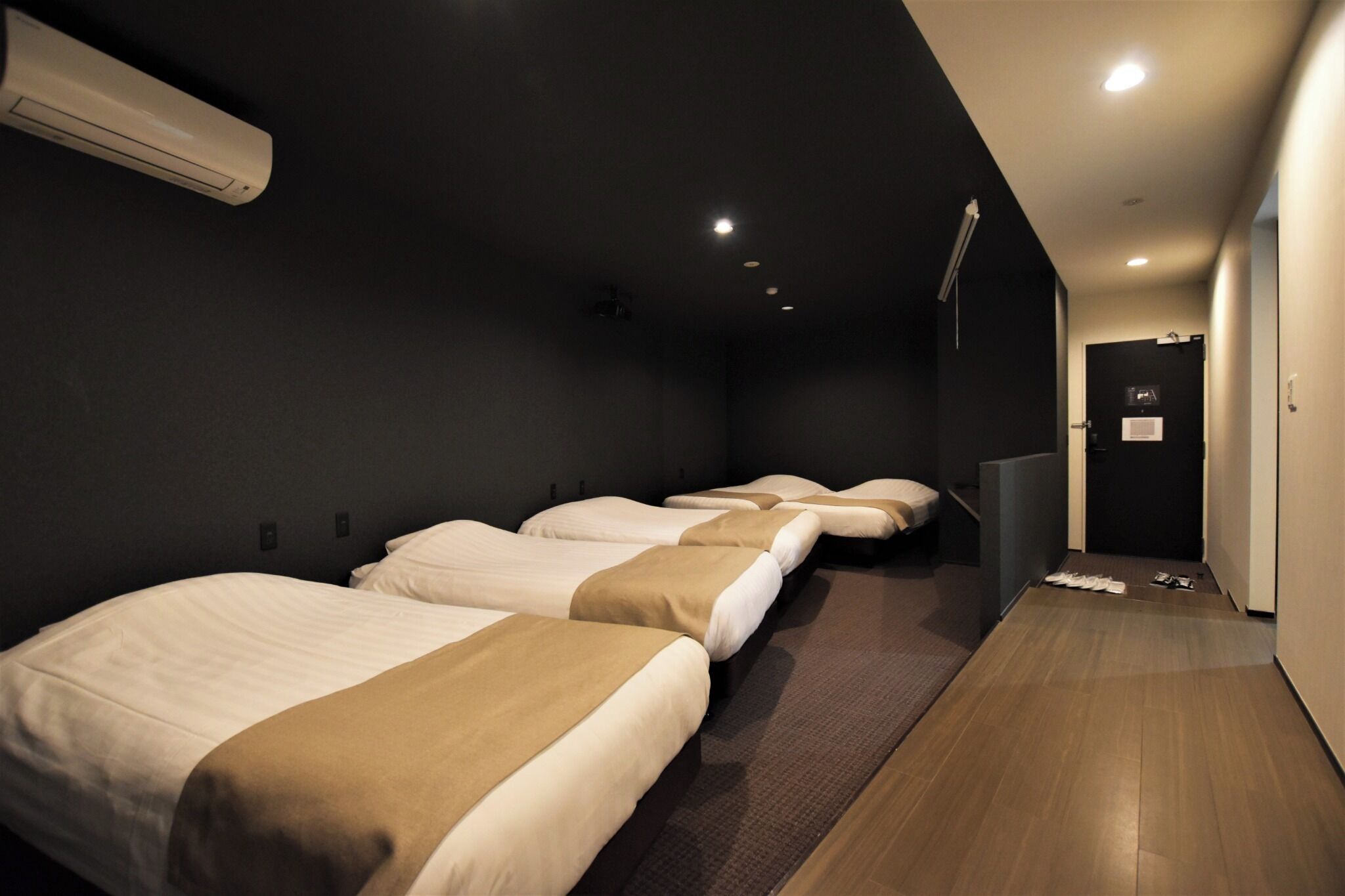 A hotel room with four beds aligned in a row, a dark wall, and a wooden floor.