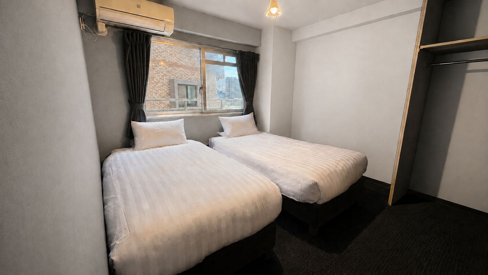 A hotel room with two beds, a window with curtains, an air conditioning unit, and a dark carpeted floor.