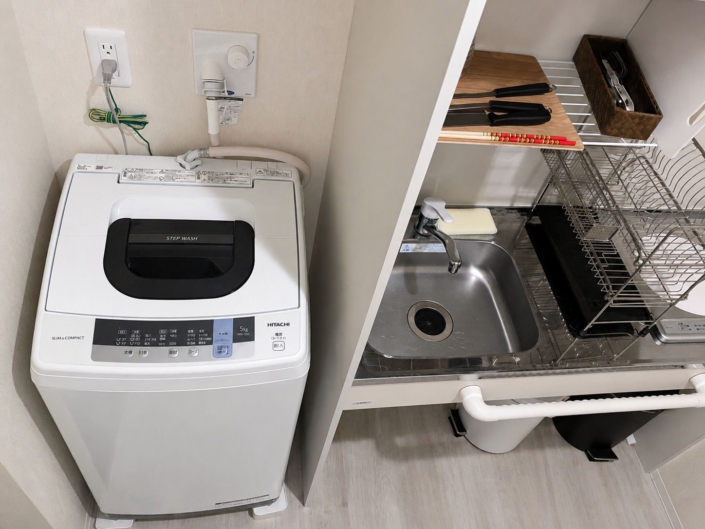 A compact washer and dryer unit with a control panel, a sink with a dish rack, and a wooden shelf with kitchen utensils.