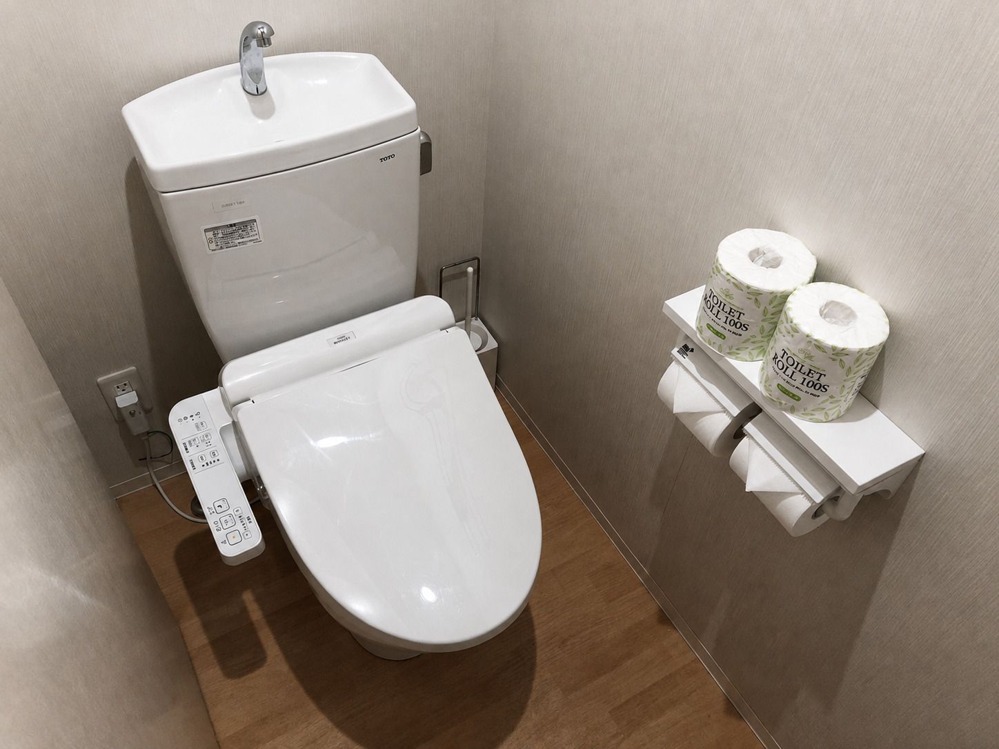 A modern toilet with a control panel, a toilet paper holder with two rolls, and a power outlet on the wall.