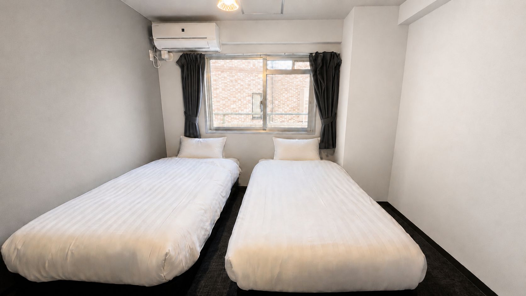 A small, clean room with two single beds, a window with curtains, and an air conditioning unit.