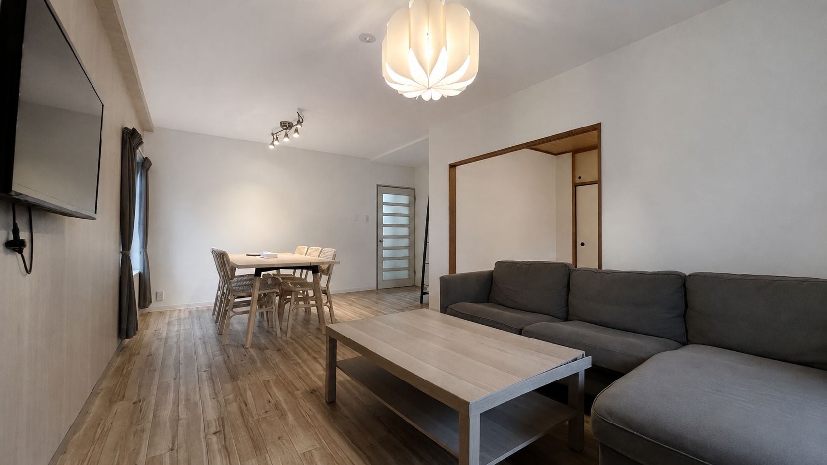 A modern living room with a sofa, a dining table with chairs, a coffee table, a TV mounted on the wall, and a ceiling light fixture.