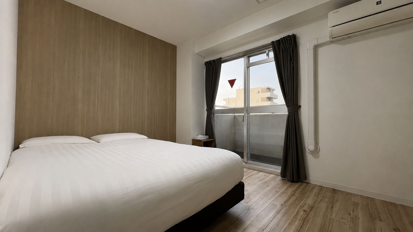 A hotel room with a bed, a nightstand, a window with curtains, and an air conditioning unit.