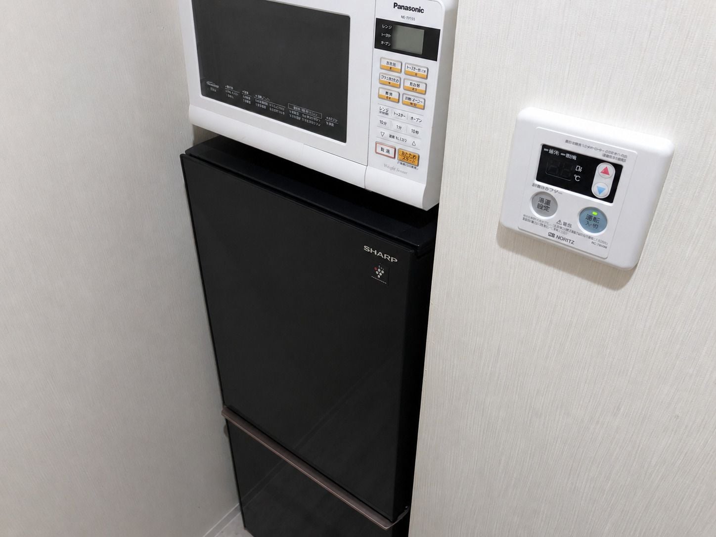 A black refrigerator with a Panasonic microwave on top, and a control panel on the wall.