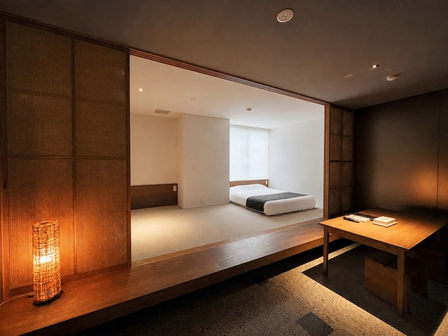 A modern room with a wooden bench, a bedside table, and a bed with a white mattress and dark bedding.