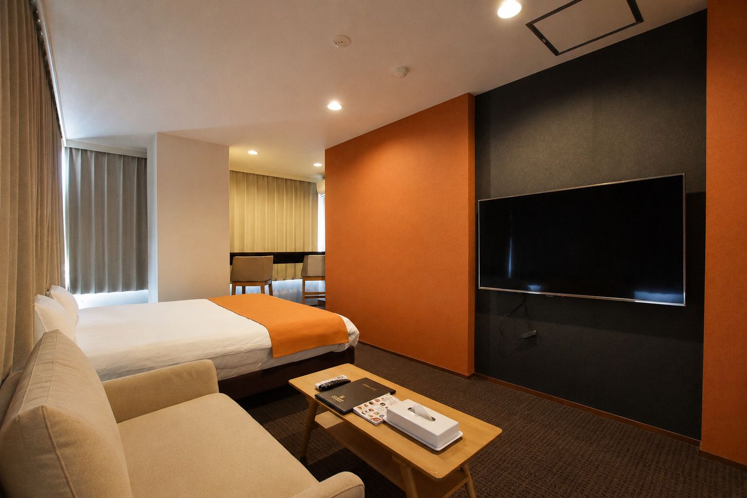 A hotel room with a bed, a sofa, a TV, a table with a remote and a tissue box, and a window with curtains.