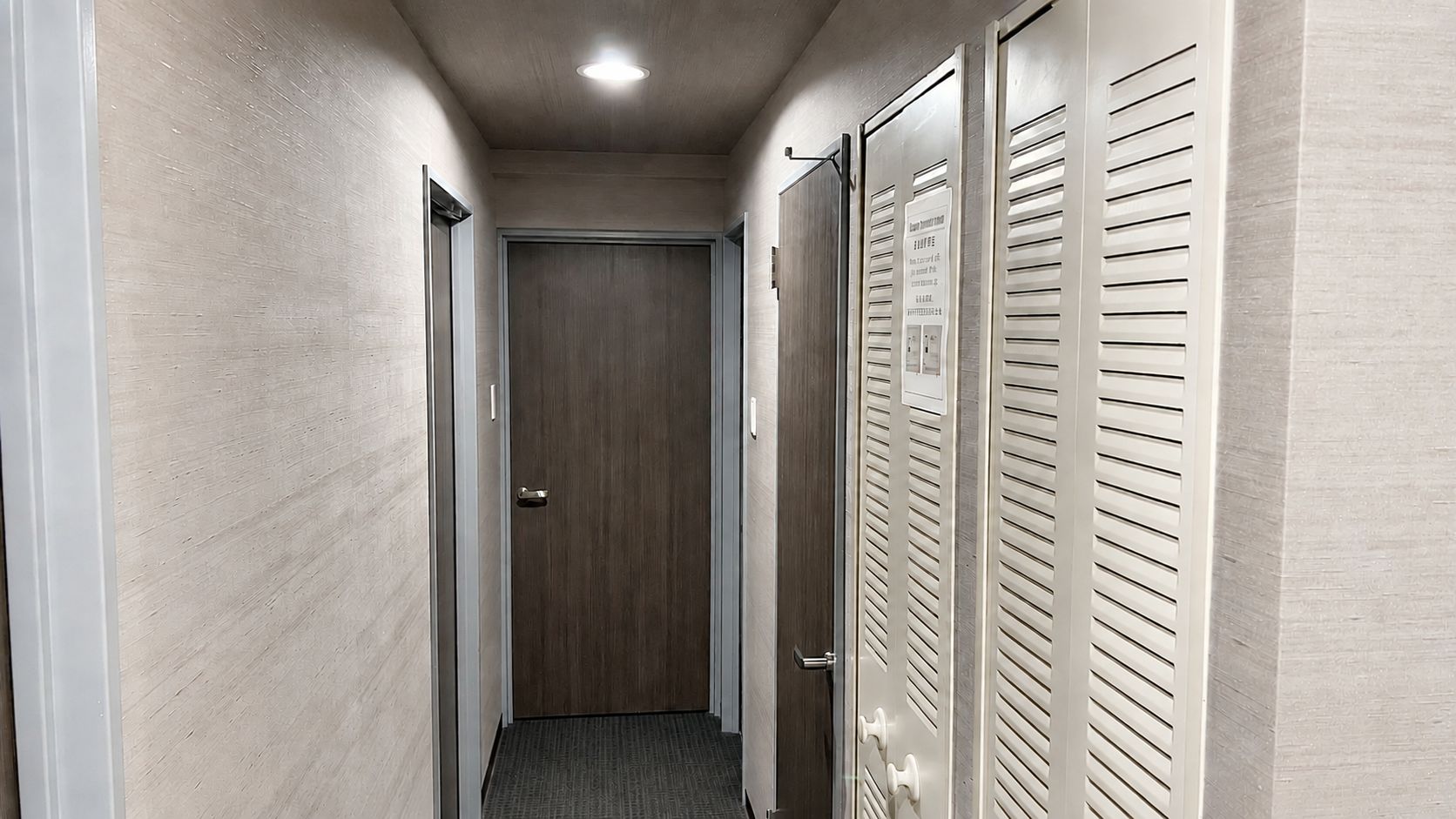 A narrow hallway with a wooden door at the end, a white door with a handle on the left, and a white door with louvered panels on the right.
