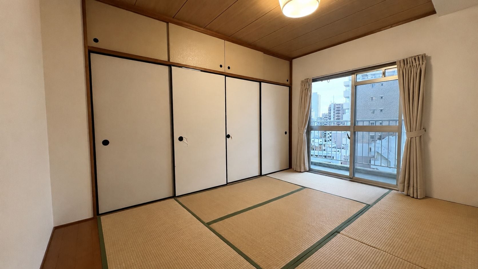 A room with tatami flooring, sliding shoji doors, a large window with curtains, and a view of buildings outside.