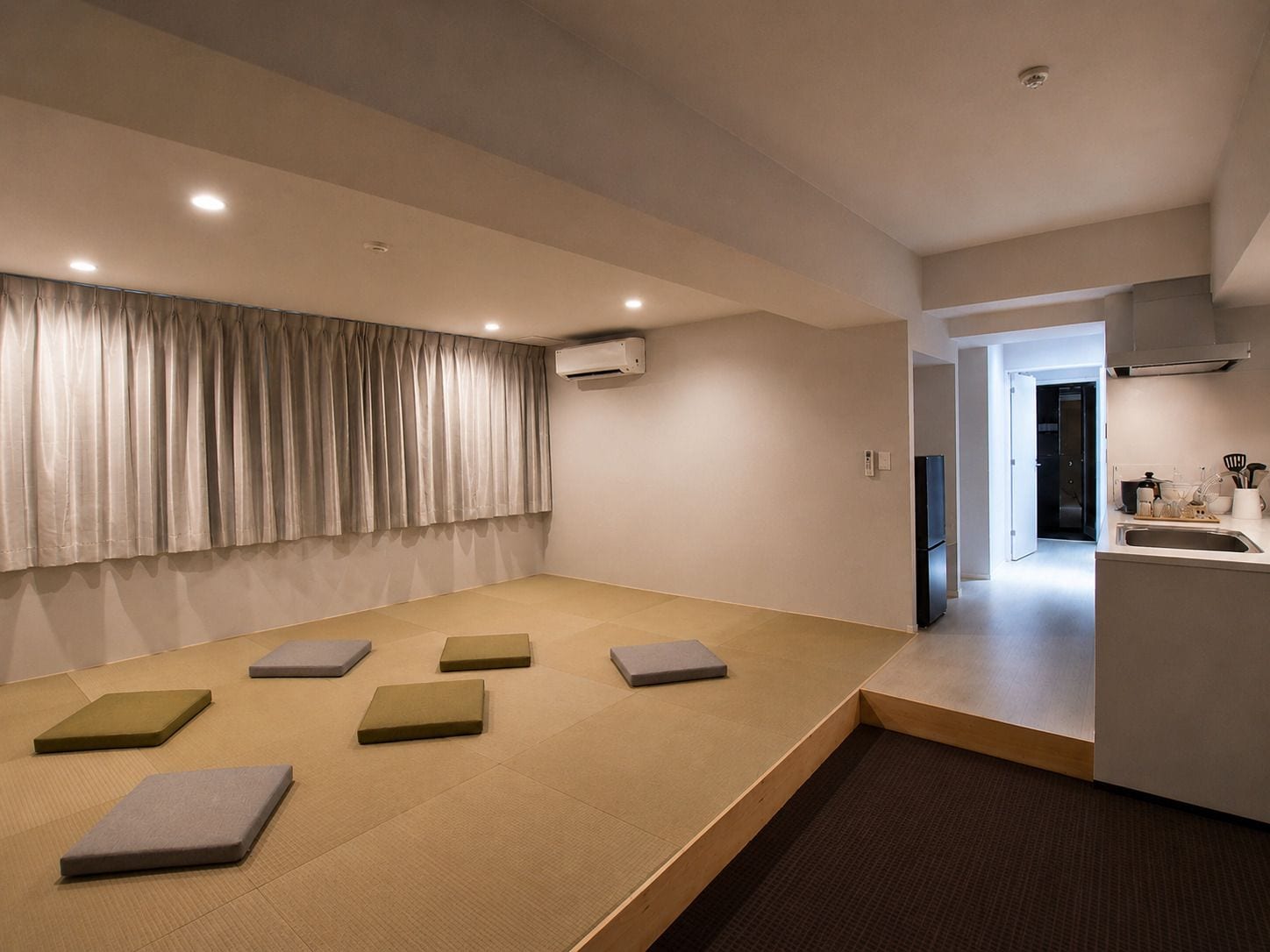 A modern living room with tatami mats, a large window with curtains, and a kitchen area visible through an open door.