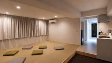 A modern living room with tatami mats, a large window with curtains, and a kitchen area visible through an open door.
