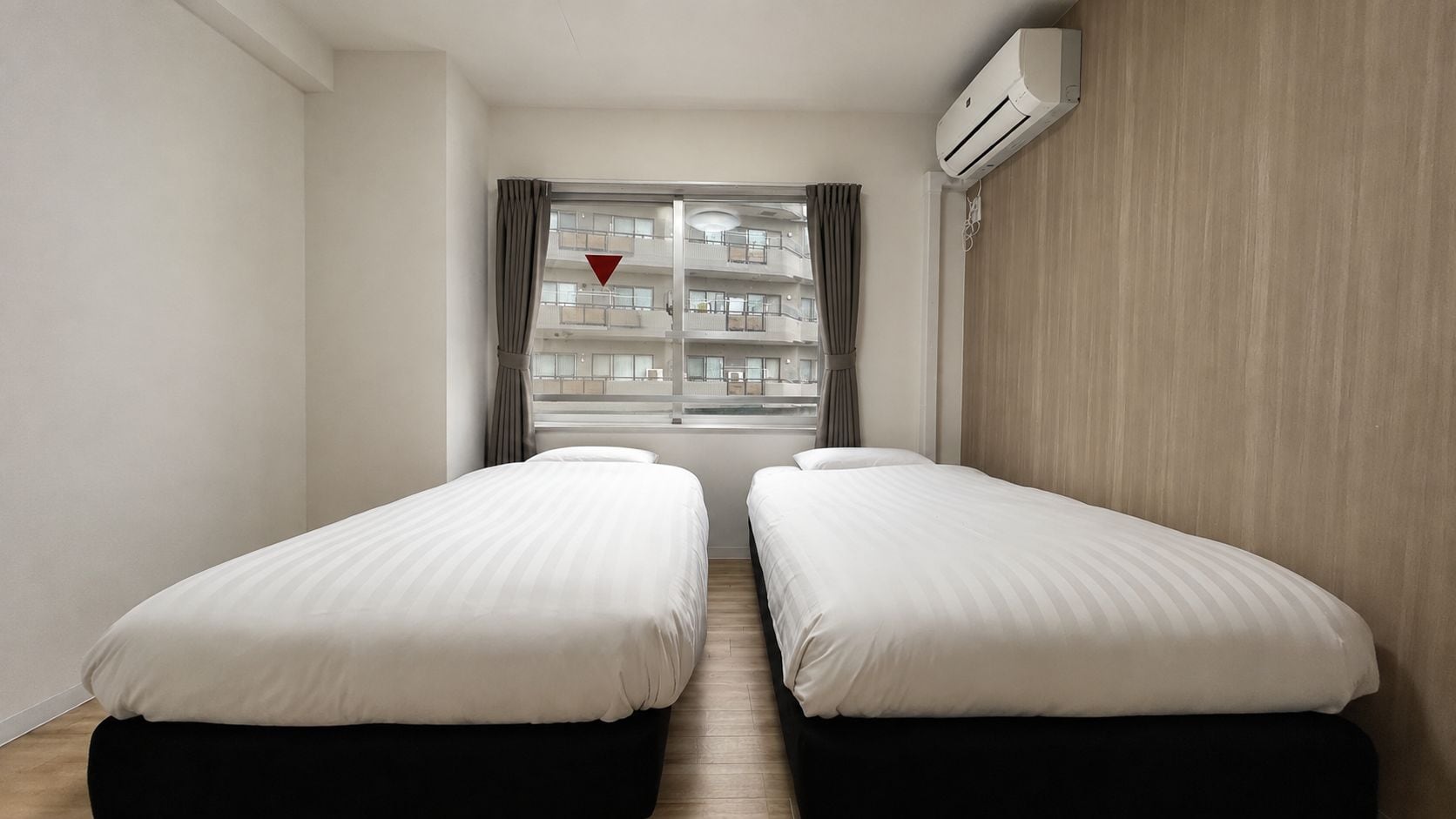 A hotel room with two beds, a window with curtains, an air conditioning unit, and a view of a building with balconies.
